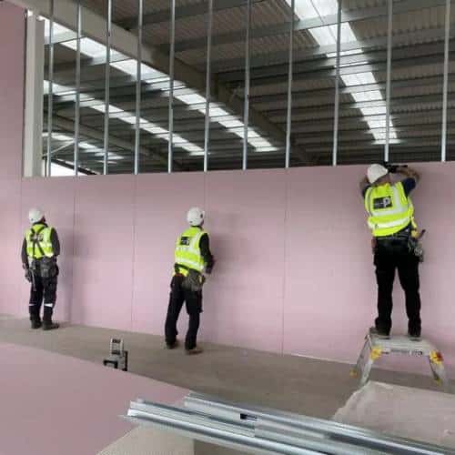 Workers in high-vis installing pink wall panels indoors.