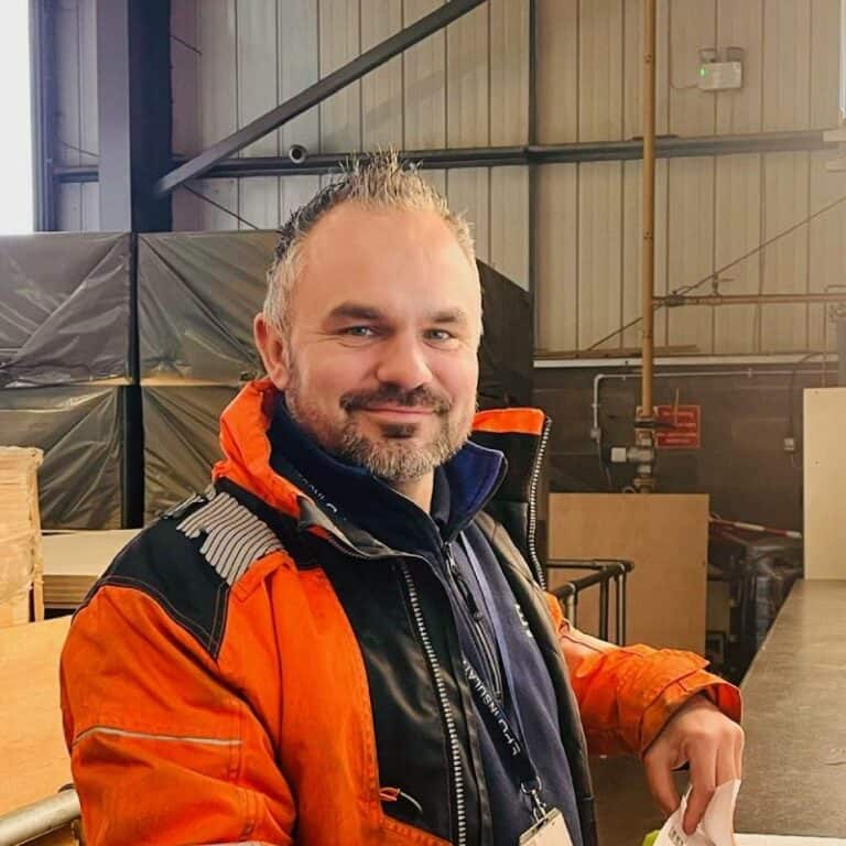 Man in orange jacket smiling in industrial warehouse.